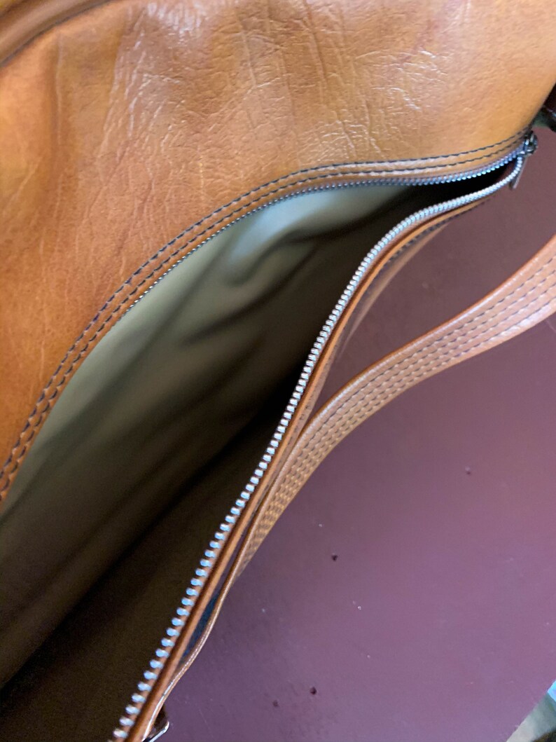 May include: A brown leather bag with a zipper closure and a brown leather strap. The bag is open and the inside is lined with a beige fabric.