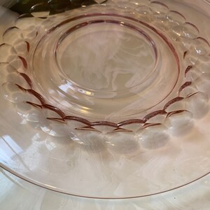 May include: A clear, pink-tinted glass plate with a raised, circular design of rounded shapes around the inner rim. The plate has a wide, flat edge and a central, slightly recessed area. The glass has a smooth, reflective surface.