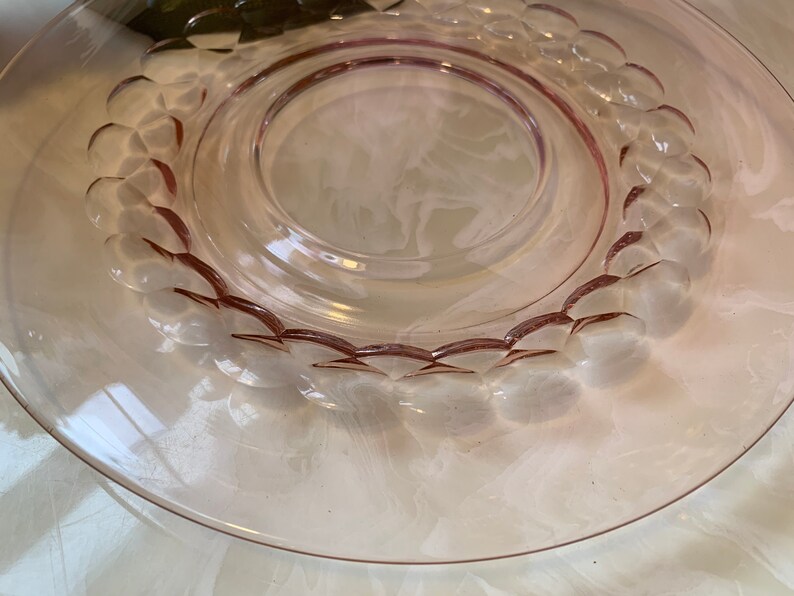 May include: A clear, pink-tinted glass plate with a raised, circular design of rounded shapes around the inner rim. The plate has a wide, flat edge and a central, slightly recessed area. The glass has a smooth, reflective surface.