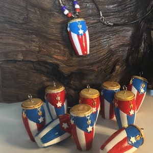 May include: A collection of ten miniature red, white, and blue Puerto Rican conga drums with gold accents. The drums are hanging from a silver hook and are arranged in a circle on a white surface. One drum is hanging from a black cord with red, white, and blue beads.