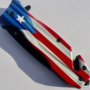 May include: A red, white, and blue folding pocket knife with a safety feature and a Puerto Rican flag design.