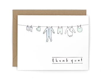 Puede incluir: Una tarjeta blanca con una línea de ropa de bebé colgada en un tendedero. La ropa incluye un gorro, calcetines, pantalones, un mono y un babero. El texto "¡gracias!" está impreso debajo del tendedero.