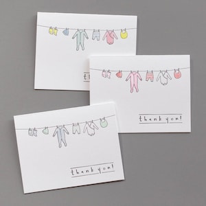 Puede incluir: Tres tarjetas de agradecimiento con una l&iacute;nea de ropa de beb&eacute; colgada en un tendedero. La ropa es de varios colores y patrones, incluyendo un body azul, un body rosa y un babero amarillo. Las tarjetas son blancas con texto negro que dice "&iexcl;gracias!"
