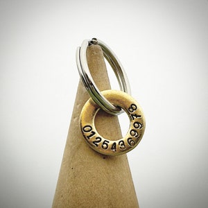 Peut inclure: Un porte-clés argenté avec un charm circulaire doré. Le charm comporte une série de chiffres sur son bord. Le porte-clés repose sur un objet brun en forme de cône sur fond blanc.