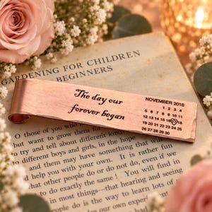 May include: A rectangular copper bookmark with a curved top edge, engraved with the text "The day our forever began" and a calendar for November 2018, featuring a heart symbol around the number 17, resting on an open book page.