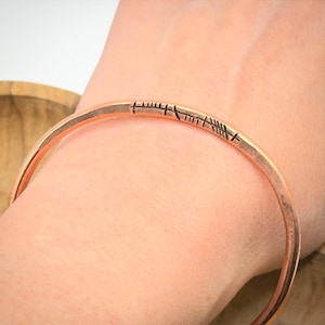 Pode incluir: Uma pulseira de cor cobre com marcações gravadas. A pulseira tem um design simples e aberto, e as marcações parecem ser uma série de linhas e traços. A pulseira é usada no pulso, e a estética geral é rústica e artesanal.