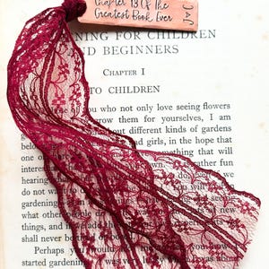 May include: A close-up of a burgundy lace ribbon with floral patterns, tied in a knot. A small wooden tag with the text "Chapter 13 Of The Greatest Book Ever" is attached. The ribbon rests on the pages of an open book.