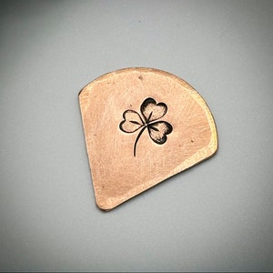 May include: A copper-coloured, irregularly shaped metal piece featuring a stamped black shamrock design. The shamrock has three heart-shaped leaves and a stem. The metal has a brushed texture and a slightly curved edge.