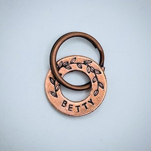 Peut inclure: Un porte-clés en métal de couleur cuivre avec une étiquette ronde portant l'inscription "BETTY" en majuscules. L'étiquette est ornée d'un motif de feuilles sur le bord. Un anneau plus grand est attaché à l'étiquette.