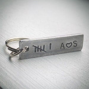 Könnte beinhalten: Ein rechteckiger Metall-Schlüsselanhänger mit silbernem Finish. Der Schlüsselanhänger hat gestempelte Markierungen, darunter die Zahl "1", die Initialen "A", ein Herzsymbol und "S". Die Zahl "5" ist durchgestrichen. Ein silberner Schlüsselring ist befestigt.