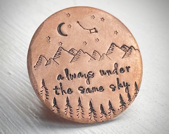 May include: A round copper-colored pendant with etched design. The design includes mountains, trees, stars, a crescent moon, and the phrase "always under the same sky".