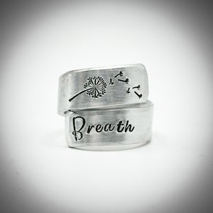 May include: A silver ring with the word "Breath" engraved on the lower band. The upper band features a dandelion design with seeds blowing away. The ring is a wrap-around style.