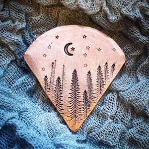 May include: Copper-toned, fan-shaped decorative item with an etched forest scene. The design includes trees, stars, and a crescent moon. The item has a handmade, rustic aesthetic.