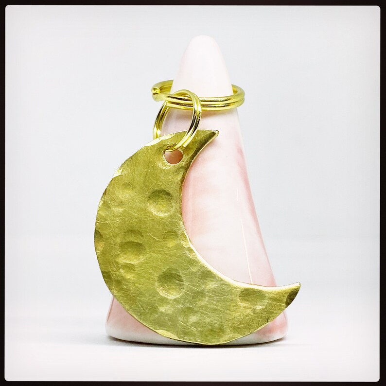 May include: A crescent moon keychain made of hammered brass, featuring a textured surface with circular indentations. The keychain has a gold-colored ring and is displayed against a pink, cone-shaped object.