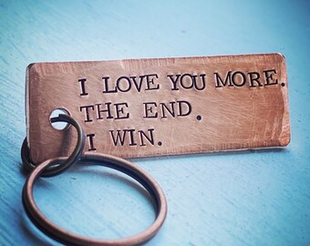 May include: A copper-colored rectangular keychain with the words "I LOVE YOU MORE. THE END. I WIN." engraved in black. The keychain is attached to a circular key ring. The background is a light blue surface.