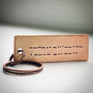 May include: A rectangular copper keyring with Morse code symbols. The keyring has a hole for the key ring and is attached to two copper-coloured rings. The Morse code is punched into the metal.