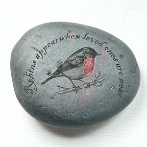 May include: A grey stone featuring a printed illustration of a robin with a red breast, perched on a branch. The text around the bird reads "Robins appear when loved ones are near."
