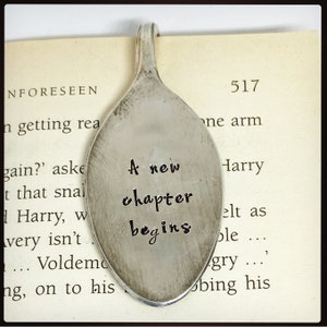 Pode incluir: Um marcador de livro de colher de prata com as palavras "A new chapter begins" estampadas na concha. O marcador de livro está colocado numa página de livro com texto visível.