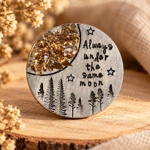 Può includere: Un disco metallico circolare color argento con una mezzaluna riempita di scaglie dorate. La parte inferiore mostra pini incisi, mentre il lato destro include tre stelle e il testo Always under the same moon.