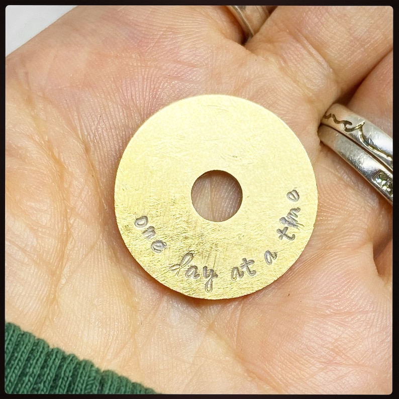 Puede incluir: Un colgante circular dorado con un agujero en el centro. El colgante est&aacute; inscrito con las palabras "one day at a time" en un estilo manuscrito. El colgante se sostiene en una mano, con un anillo de plata visible en el fondo.