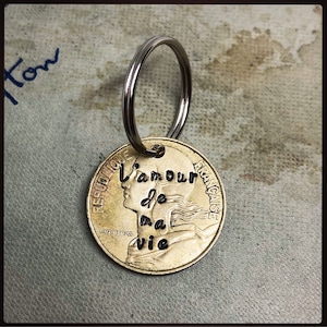 May include: A gold-coloured coin keyring with the words "L'amour de ma vie" stamped on it. The coin features a profile of a woman and the words "REPUBLIQUE FRANCAISE". A silver-coloured key ring is attached to the coin.