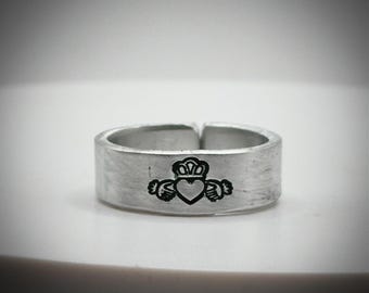 May include: A silver ring featuring a Claddagh design. The Claddagh symbol, with a heart, crown, and hands, is etched in black on the ring's band. The ring has a brushed metal finish and an adjustable design.