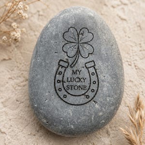 May include: A smooth, oval-shaped gray stone featuring a black engraved design of a four-leaf clover positioned above a horseshoe. The center of the horseshoe contains the text MY LUCKY STONE in clear, capitalized black lettering.