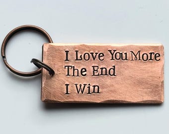 May include: A rectangular copper keychain with the words "I Love You More The End I Win" engraved in a bold, sans-serif font. The keychain has a textured surface and a bronze-colored key ring.
