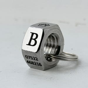 Peut inclure: Écrou métallique argenté avec un porte-clés. L'écrou porte la lettre "B", une empreinte de patte et les chiffres "07522" et "468216". Il s'agit probablement d'un porte-clés ou d'un pendentif.