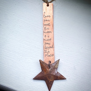 May include: A copper-colored metal bookmark with the engraved text "i love you past the moon & i miss you beyond the stars." A rusty star-shaped charm hangs below the bookmark, attached by a metal ring. The bookmark is suspended by a natural twine.