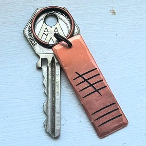 Puede incluir: Una llave con una etiqueta de cobre. La etiqueta rectangular tiene seis líneas negras grabadas en el cobre. La llave tiene un anillo de cobre y un lazo negro. La llave es plateada y tiene un diseño único.