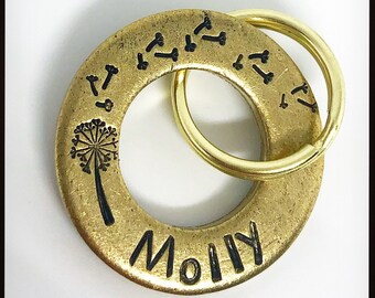May include: A gold-colored metal keychain with a circular pendant. The pendant features the name "Molly" in black lettering, a dandelion design, and several small bone-shaped designs. A gold-colored ring holds the pendant.
