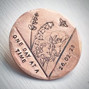 Può includere: Un disco metallico rotondo color rame con disegni floreali incisi, tra cui lavanda, mughetto, un papavero e un'ape. Il testo "ONE DAY AT A TIME" e la data "26.02.23" sono anch'essi incisi.