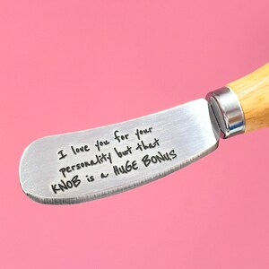 May include: A butter knife with a stainless steel blade and a wooden handle. The blade is engraved with the text: "I love you for your personality but that KNOB is a HUGE BONUS" against a pink backdrop.