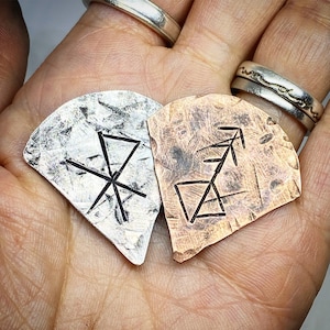 May include: Two metal pendants with etched symbols. One is silver with a starburst design, the other is copper-toned with a geometric arrow-like symbol. Both are held in a hand, showcasing their textures and details.
