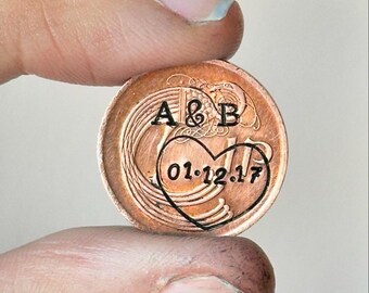 May include: A copper coin featuring the initials "A & B" and the date "01.12.17" within a heart. The coin is held up, displaying the engraved design and the warm copper tone.