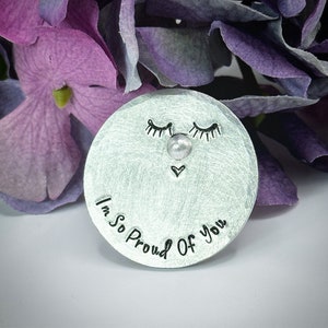 Può includere: Un ricordo rotondo in metallo argentato con la frase "I'm So Proud Of You". Presenta un design a forma di viso con occhi chiusi, un naso perlato e un piccolo cuore. Fiori viola e lavanda sullo sfondo.