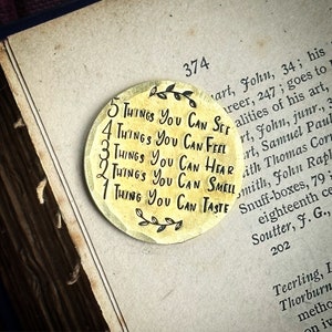 May include: A circular, gold-toned metal pendant inscribed with the words "5 Things You Can See, 4 Things You Can Feel, 3 Things You Can Hear, 2 Things You Can Smell, 1 Thing You Can Taste". The pendant rests on an open book.