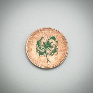 Puede incluir: Una moneda de un penique de color cobre con un diseño de trébol de cuatro hojas verde en el centro. La moneda tiene las palabras "ONE PENNY" alrededor del borde.