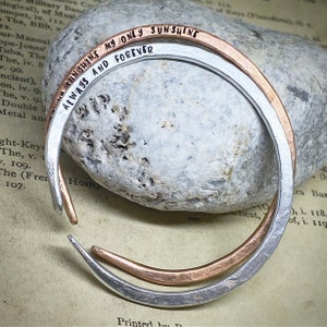 May include: Two metal bangles, one silver and one copper, are shown. The silver bangle has the words "ALWAYS AND FOREVER" stamped on it. The copper bangle has the words "MY ONLY SUNSHINE" stamped on it. The bangles are on a stone.