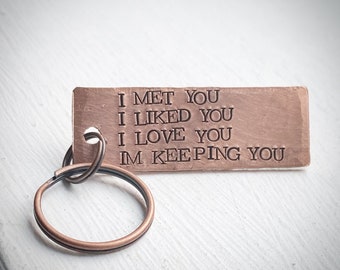 May include: A copper-colored rectangular keychain with a key ring. The keychain has the stamped text: "I MET YOU, I LIKED YOU, I LOVE YOU, IM KEEPING YOU". The key ring is made of a similar copper-colored metal.