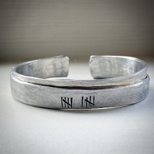 Può includere: Un bracciale a polsino color argento con finitura in metallo spazzolato. Il bracciale presenta segni di conteggio incisi, probabilmente per un conteggio o una data. Il design aperto ne facilita l'indossamento.