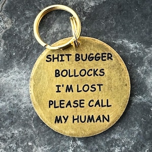 May include: A circular, brass pet tag with a gold-tone ring. The tag features the phrases "SHIT BUGGER BOLLOCKS I'M LOST PLEASE CALL MY HUMAN" in black, sans-serif lettering.