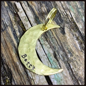 May include: A hammered brass crescent moon pendant with a gold-colored loop. The word "Betty" is stamped on the moon's surface. The pendant is displayed on a weathered wooden background.