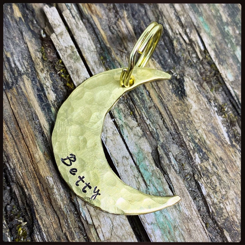 May include: A hammered brass crescent moon pendant with a gold-colored loop. The word "Betty" is stamped on the moon's surface. The pendant is displayed on a weathered wooden background.