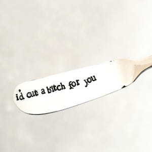 May include: A stainless steel butter knife with the phrase "i'd cut a bitch for you" stamped in black, typewriter-style font. The knife has a rounded tip and a long, flat blade.