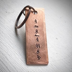 May include: A copper-coloured rectangular keyring with a bronze key ring. The keyring has stamped symbols and a small bird illustration. The background is a light, textured surface.
