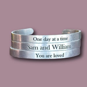 Könnte beinhalten: Silbernes Metallarmband mit drei übereinanderliegenden Bändern. Das obere Band lautet "One day at a time", das mittlere Band "Sam and William" und das untere Band "You are loved".