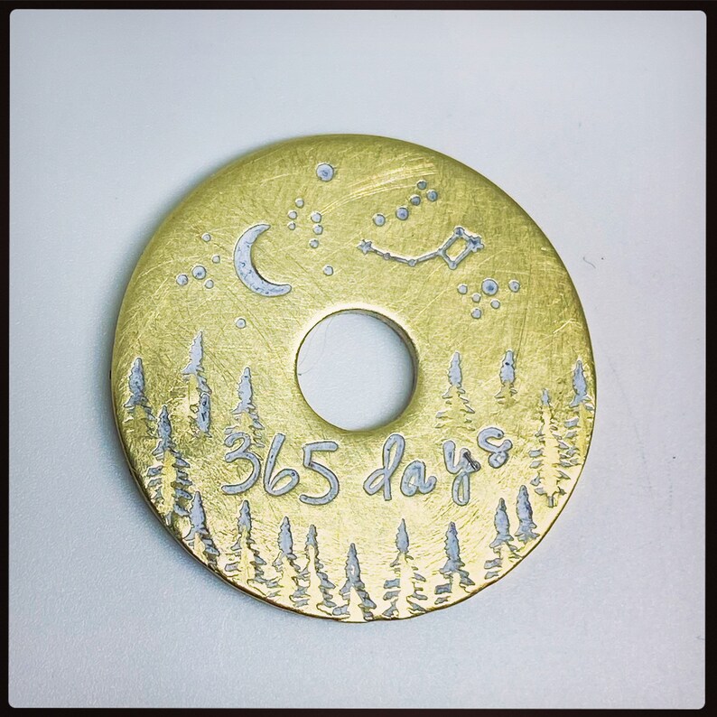 Puede incluir: Un colgante circular dorado con un agujero central. El colgante presenta una escena de bosque en la parte inferior, una luna creciente, estrellas y el texto "365 days". El dise&ntilde;o est&aacute; grabado en plata sobre el fondo dorado.