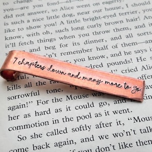 May include: A copper bookmark with a curved end, featuring the hand-stamped black text 7 chapters down and many more to go. The metal has a hammered texture and a warm, metallic finish, resting against a page of printed book text.